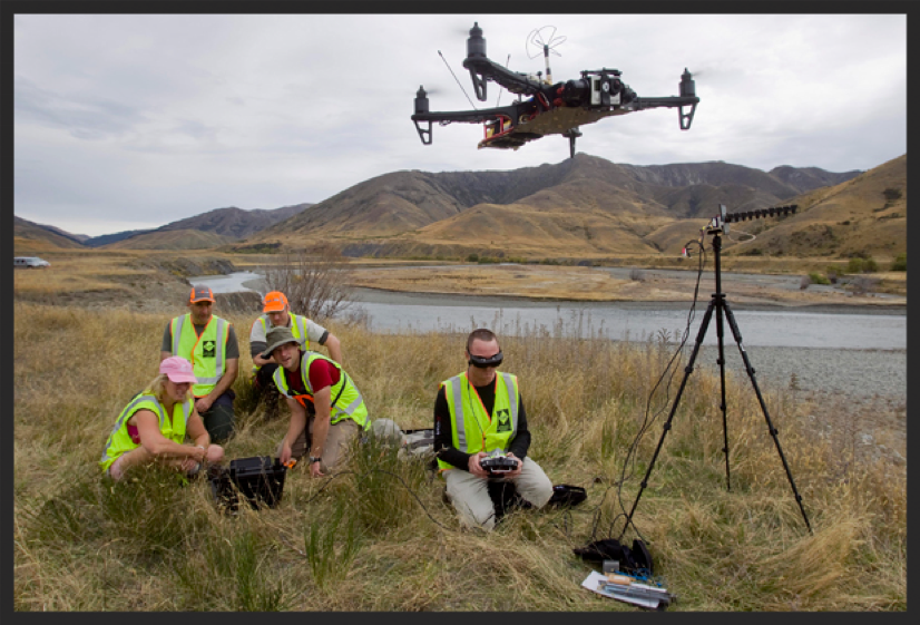 A field team in high-visibility vests flying a drone beside a river valley