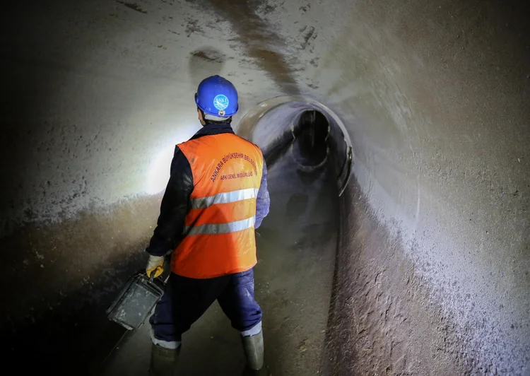 A sewer worker in a hard hat and high-visibility vest walking through a corroded concrete tunnel